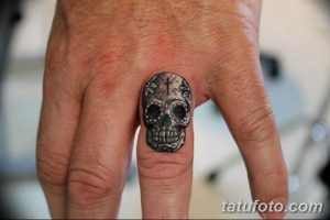Фото тату маленького черепа с крестом на пальце руки. Четкий и лаконичный рисунок в черно-серых тонах.