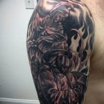 Фото тату на предплечье с изображением пожарного в полном обмундировании, окруженного языками пламени. Детализированный рисунок в черно-серых тонах.