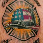 Фото тату шлема пожарного, украшенного флагом США, с надписями 'Fallen Brothers' и 'Never Forgotten'. Патриотичный рисунок татуировки.