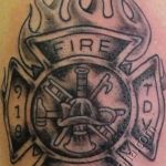 Фото тату пожарного шлема с крестом, пламенем и надписями 'FIRE' и 'RESCUE', выполненное в черно-белом стиле.