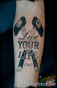 Фото тату с надписью 'Live Your Life' и изображением двух пистолетов, выполненное в черном цвете. Стильная и вдохновляющая татуировка на предплечье.