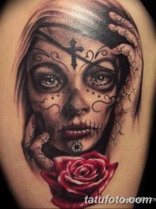 Фото тату: татуировка в стиле Sugar Skull с изображением черепа, украшенного узорами, с розой и крестом, выполненная на плече.