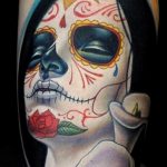 Фото тату: красочная татуировка в стиле Sugar Skull (Калавера) с изображением черепа, украшенного цветами и узорами, и красной розой на предплечье.