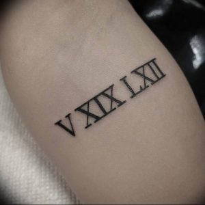 Фото тату: черные римские цифры 'V XIX LXII' на предплечье. Минималистичный дизайн, четкие линии, небольшое изображение.