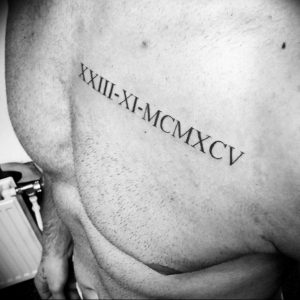 Фото тату на груди мужчины: римские цифры 'XXIII-XI-MCMXCV' выполнены черными чернилами, линейный дизайн.