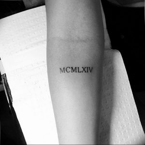 Черно-белое фото татуировки с римскими цифрами MCMXLIV на запястье. Минималистичный дизайн, тонкие линии, небольшие размеры.