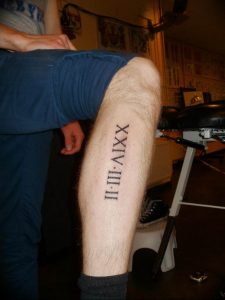 Фото тату: черные римские цифры 'II-III-XIV-III' на икре мужчины. Простой и лаконичный дизайн, четкие линии.