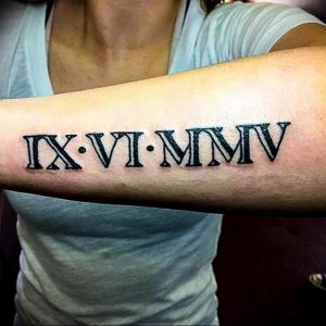 Фото тату: черные римские цифры 'IX-VI-MMMV' на внутренней стороне предплечья женщины. Элегантный минималистичный дизайн, четкие линии.