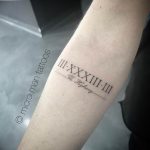 Черно-белое фото микро татуировки на предплечье с римскими цифрами III-XXXIIII-III и надписью "The Highway". Тонкие линии, минималистичный дизайн.