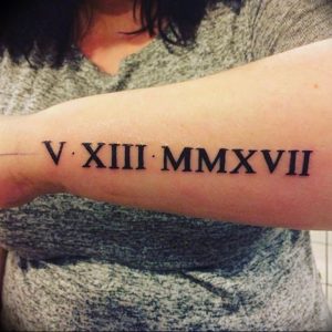 Черно-белое фото татуировки на предплечье с римскими цифрами V-XIII-MMXVII. Минималистичный дизайн, четкие линии, символ важной даты.
