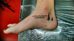 Фото татуировки на лодыжке с изображением римских цифр 'V.XVIII.MCMXLVIII X.XXI.MMVII' и двух небольших елей.