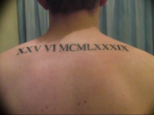 Фото тату: большая надпись римскими цифрами 'XXV VI MCMLXXXIX' на спине мужчины. Черный цвет, изогнутая форма, минималистичный дизайн.