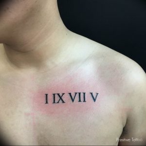 Фото тату: черные римские цифры 'I IX VII V' на груди мужчины, с легким эффектом покраснения кожи вокруг. Минималистичный дизайн, реалистичный эффект.