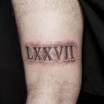 Фото тату: римские цифры 'LXXVII' на предплечье мужчины, стилизованные под шрам. Черный цвет, реалистичный эффект, брутальный дизайн.