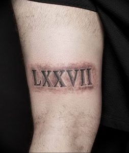 Фото тату: римские цифры 'LXXVII' на предплечье мужчины, стилизованные под шрам. Черный цвет, реалистичный эффект, брутальный дизайн.