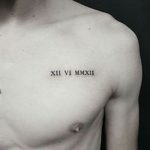 Фото тату: черные римские цифры 'XII VI MMXXII' на груди молодого человека. Минималистичный дизайн, четкие линии, небольшое изображение.