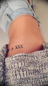 Фото тату: небольшая татуировка римскими цифрами 'X.IV.XXX' на женском боку, выполненная в черном цвете. Изящный и лаконичный рисунок.