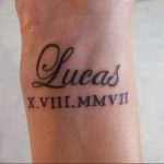 Фото татуировки на запястье. Изображено имя 'Lucas' и дата рождения в римских цифрах. Черно-белый минималистичный дизайн.