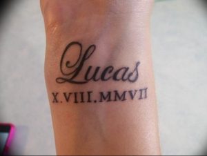 Фото татуировки на запястье. Изображено имя 'Lucas' и дата рождения в римских цифрах. Черно-белый минималистичный дизайн.