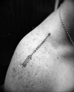 Фото татуировки на плече. Изображена тонкая стрела с римскими цифрами вдоль тела стрелы. Черно-белый минималистичный дизайн.
