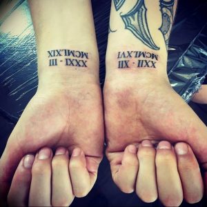 Фото парных тату на запястьях с черными римскими цифрами XIX.III.MCMXLIX и XVI.IX.MCMXLII. Минималистичный дизайн, небольшие размеры, символическое значение.