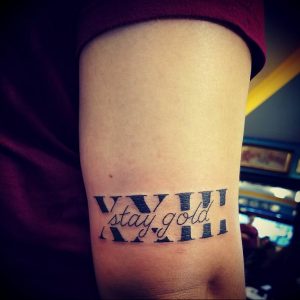 Фото тату на предплечье с черными римскими цифрами V.VII.II и надписью "stay gold". Сочетание цифр и текста, оригинальный дизайн.