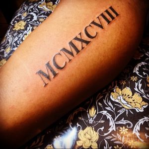Фото тату на руке с черными римскими цифрами MCMXCVIII. Длинная горизонтальная линия, четкий шрифт, минималистичный дизайн.