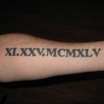 Фото тату на предплечье с черными римскими цифрами XI.XXV.MCMXLV. Горизонтальное расположение, четкий шрифт, минималистичный дизайн.