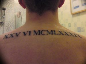 Фото тату на спине, в области шеи и лопаток, с черными римскими цифрами XXV VI MCMLXXXIX. Изогнутая линия, минималистичный дизайн.