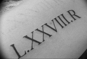 Фотография простой татуировки с римскими цифрами "LXXVIII.R" на коже, выполненной в черном цвете.