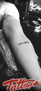 Фото тату: черные римские цифры 'XXV I MMVII' на бедре девушки. Изящный и лаконичный дизайн, подчеркивает женственность.