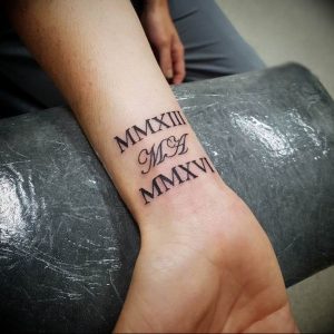 Фото тату на запястье с римскими цифрами 'MMXIII', инициалами 'M.H.' и 'MMXVI', выполненной черными чернилами. Элегантный и минималистичный дизайн.