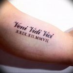 Фото тату на предплечье с надписью 'Veni Vidi Vici' и римскими цифрами, выполненной черными чернилами. Идеально для любителей истории и латыни.