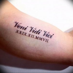 Фото тату на предплечье с надписью 'Veni Vidi Vici' и римскими цифрами, выполненной черными чернилами. Идеально для любителей истории и латыни.