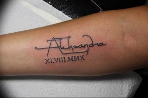 Фото тату: На руке изображено имя "Alexandra" и дата в римских цифрах XI.VIII.MMX. Стильный и минималистичный дизайн.
