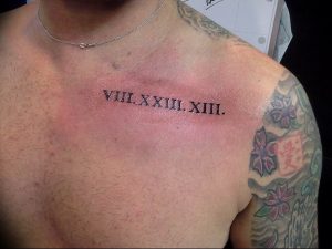Фото тату на груди у мужчины: горизонтальная линия римских цифр, выполненная черным цветом, с татуировкой на плече.