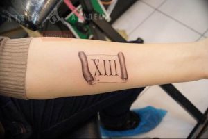 Фото татуировки на руке в виде свитка с римскими цифрами 'XIII'. Детализированный рисунок, выполненный в технике реализма.