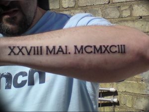 Фото тату: длинная фраза, написанная римскими цифрами, расположена на предплечье мужчины. Классический и стильный рисунок татуировки.