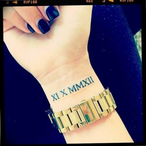 Фото тату: Татуировка с римскими цифрами XI X MMXII на запястье девушки. Черный цвет, тонкие линии, изящный дизайн, золотые часы.
