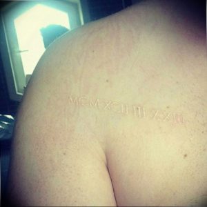 Фото тату: белые римские цифры 'MCMXCII III LXIII' на спине человека, выполненные в минималистичном стиле.