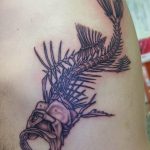 Фото тату: черно-белый рисунок скелета рыбы, выполненный в тонких линиях на руке. Элегантный дизайн и минималистичный стиль.
