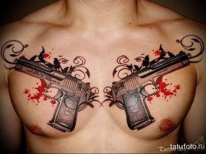 Фото тату: два скрещенных пистолета с красными брызгами, имитирующими кровь, на мужской груди. Рисунок татуировки выполнен в черно-серых тонах с добавлением красного цвета.