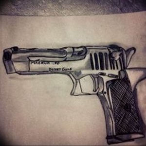 Фото тату реалистичного пистолета Desert Eagle с детальной проработкой текстуры и надписями. Черно-серый рисунок.