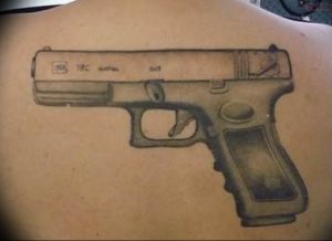 Фото тату реалистичного пистолета Glock на спине мужчины. Черно-серый рисунок с детальной проработкой механизма и надписями.