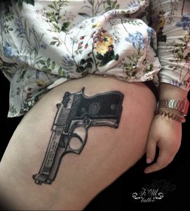 Фото тату: реалистичный рисунок револьвера на женском бедре, выполненный в черно-серых тонах. Детализированный эскиз татуировки.
