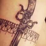 Фото татуировки старинного пистолета с орнаментом на спине. Детализированный рисунок татуировки в черно-серых тонах.