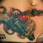Фото татуировки револьвера, пронзающего сердце, в стиле олдскул. Яркий рисунок татуировки с использованием красного и синего цветов.