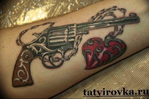 Фото тату пистолета и сердца, соединенных цепью на руке. Черно-красные тона, детализированная проработка металла и узоров.
