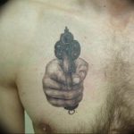 Фото тату: реалистичный рисунок руки, держащей пистолет, выполненный на груди. Детализированная татуировка в черно-серых тонах.