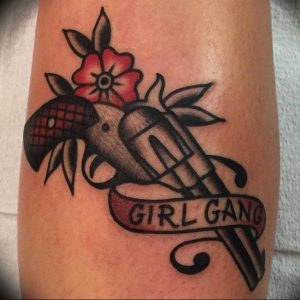 Фото татуировки револьвера с розой и надписью "Girl Gang" в стиле олдскул, выполненной в ярких цветах.